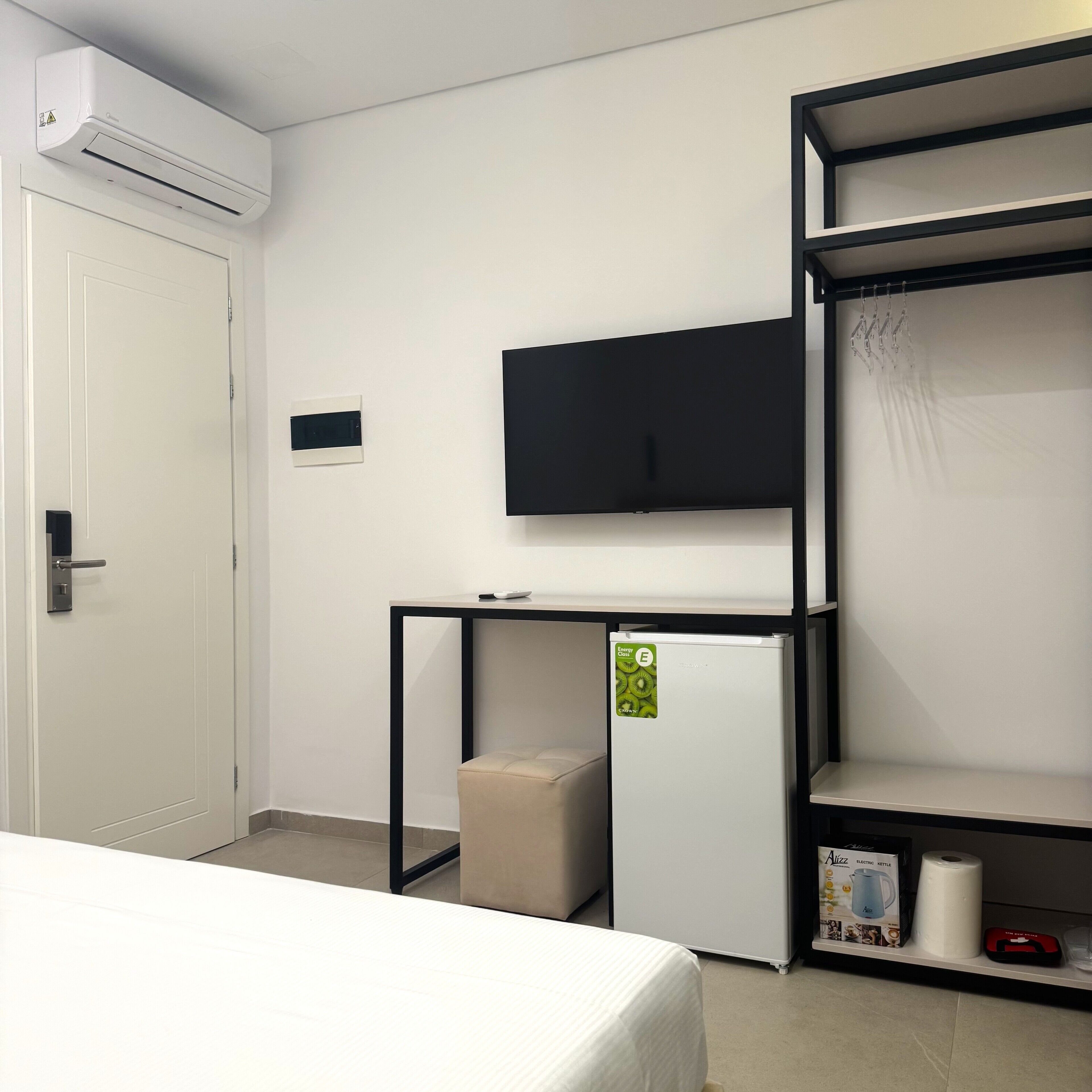 A hotel room with a bed, a small kitchenette, a TV mounted on the wall, and a shelf with a kettle and other items.