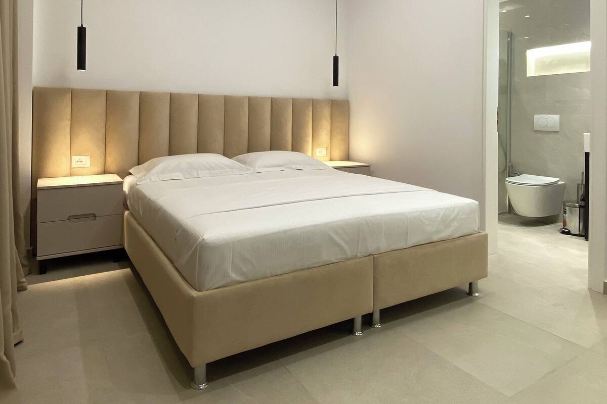 A modern bedroom with a large bed, bedside tables, and a bathroom visible through a glass door.