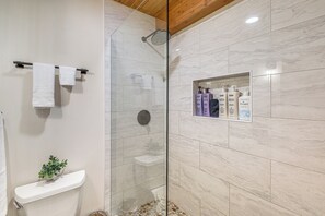 A modern bathroom with a glass-enclosed shower, a toilet with a plant, and a shelf with toiletries. - Whitefish Lake Couples Escape w/ Community Perks! (Whitefish)