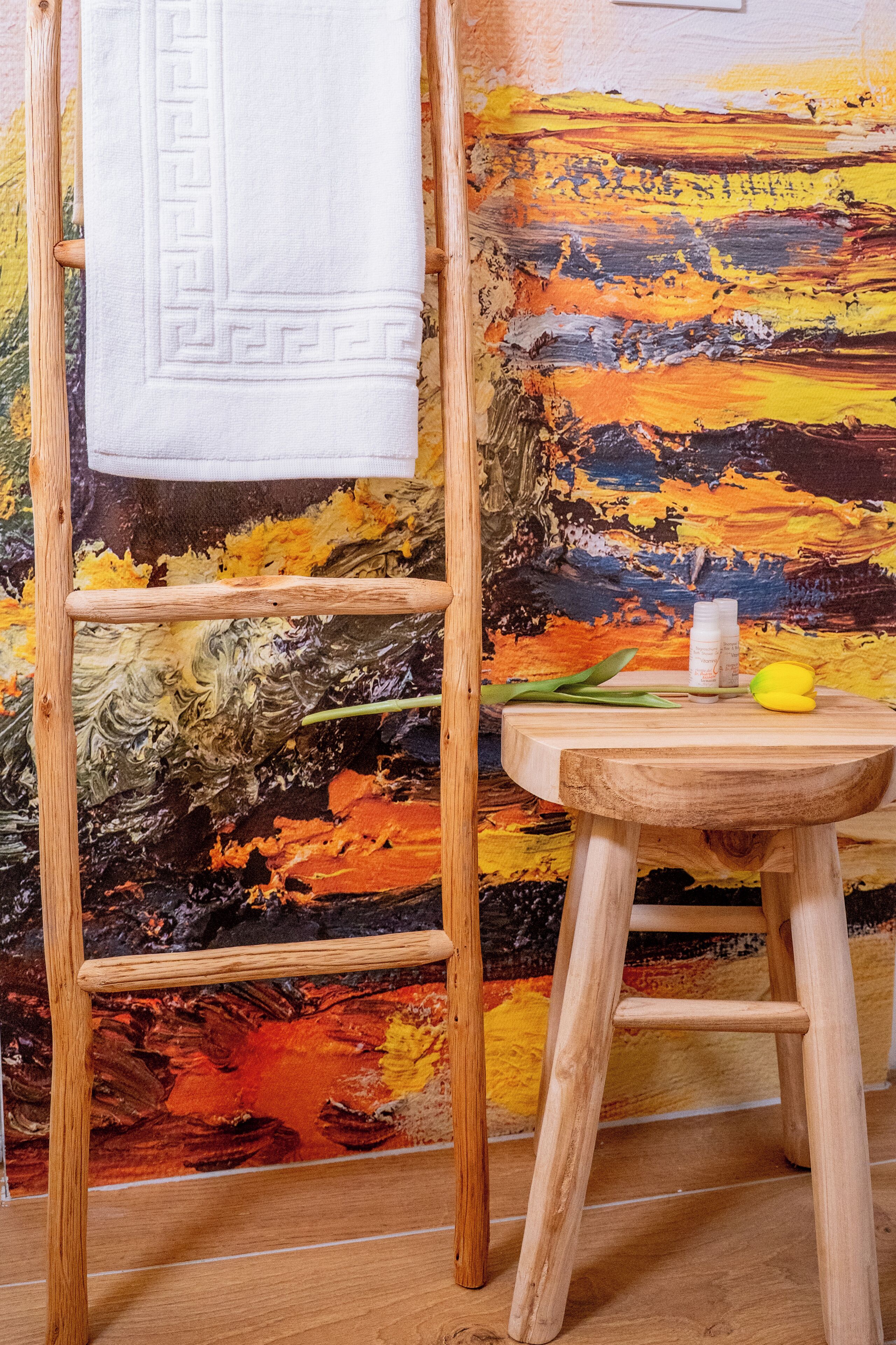 A wooden ladder leaning against a colorful abstract painting, a small wooden stool with a bottle and a lemon on it, and a white towel hanging on the ladder.
