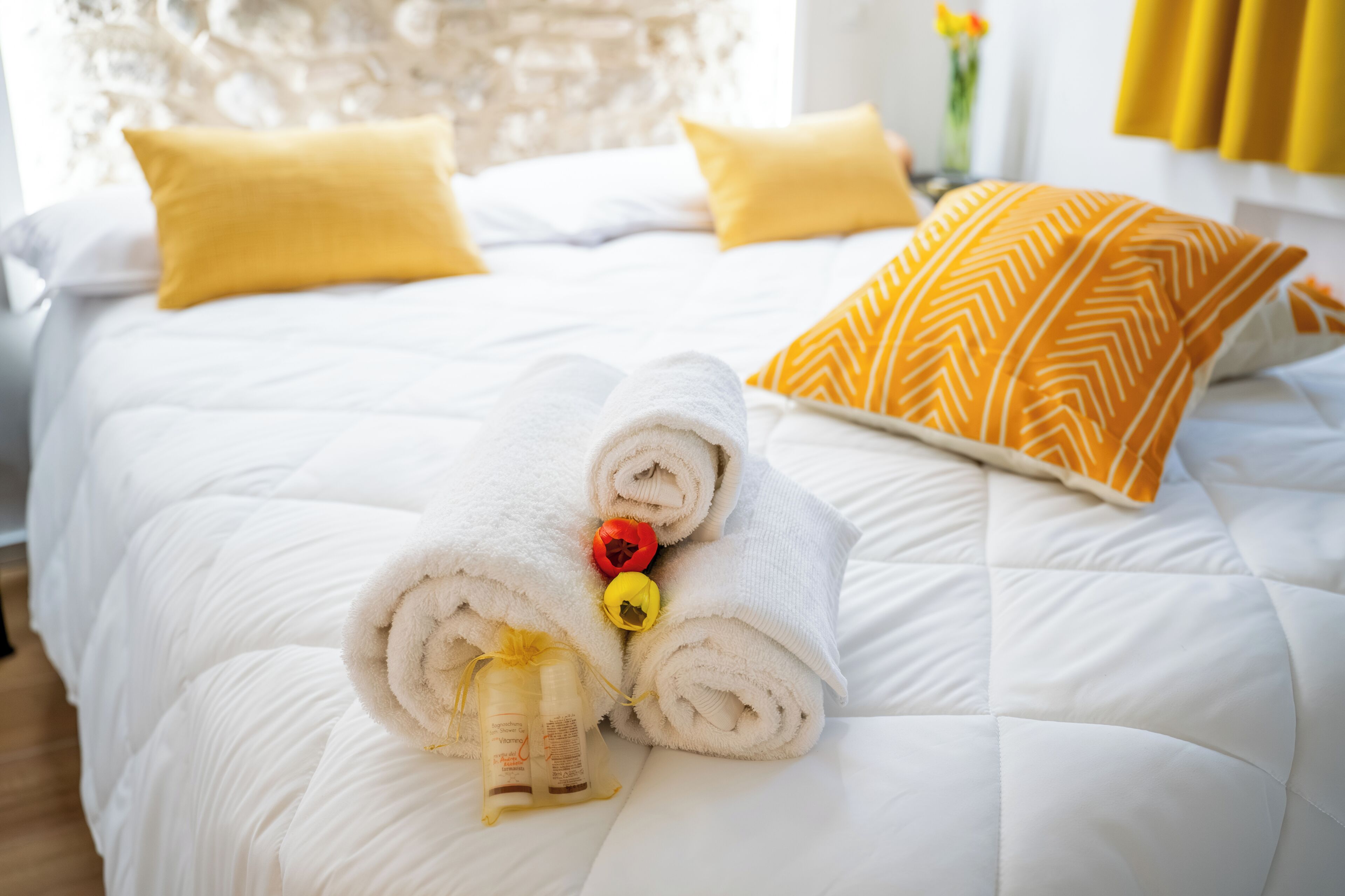Een net opgemaakt bed met opgerolde handdoeken en een klein cadeautje, gele kussens en een gestreept kussen op een wit dekbed.