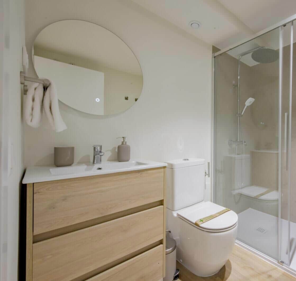 Une salle de bain moderne avec un meuble-lavabo en bois, un miroir rond, des toilettes et une douche à parois de verre.