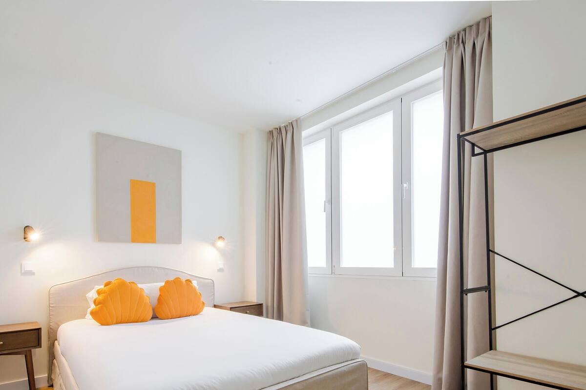 Un dormitorio con cama, mesitas de noche, una ventana y una lámpara fijada en la pared.