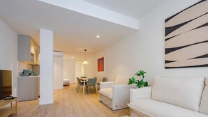 Un salon moderne avec un canapé, un coin repas avec des chaises, un téléviseur et une cuisine en arrière-plan.