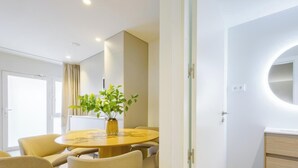 Une chambre d’hôtel moderne avec une table de salle à manger ronde, des chaises, une plante en pot, un miroir rond et une coiffeuse au design minimaliste.