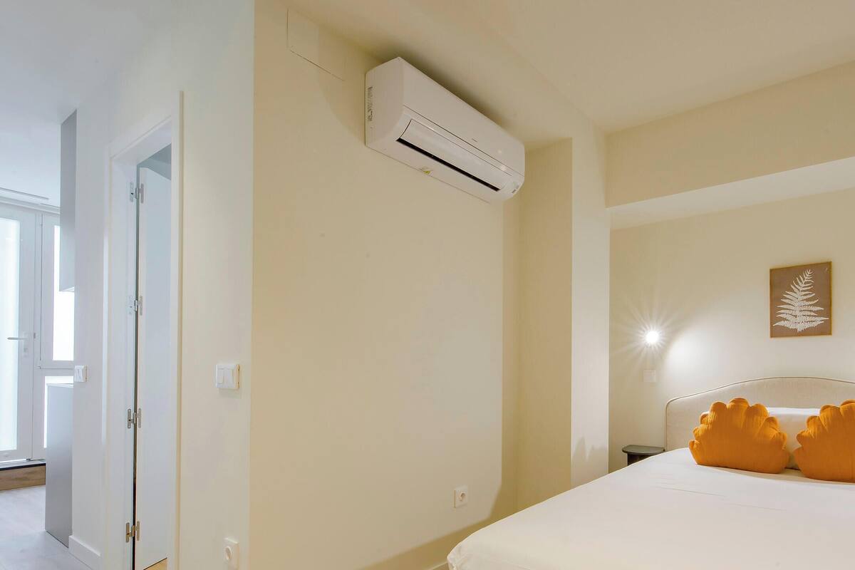 Una habitación de hotel con cama, unidad de aire acondicionado, mesita de noche y lámpara de pared.