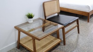 Een kamer met een bed, een tafel met glazen blad en een geweven stoel, een kleine kamerplant en een kunstwerk aan de muur.