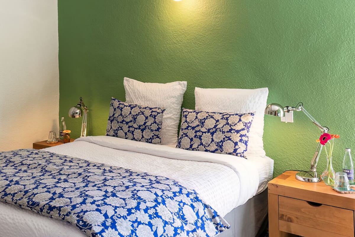 Un dormitorio con pared verde, cama con colcha floral azul y blanca, almohadas blancas, mesita de noche con lámpara y una mesita auxiliar con botella y vasos.