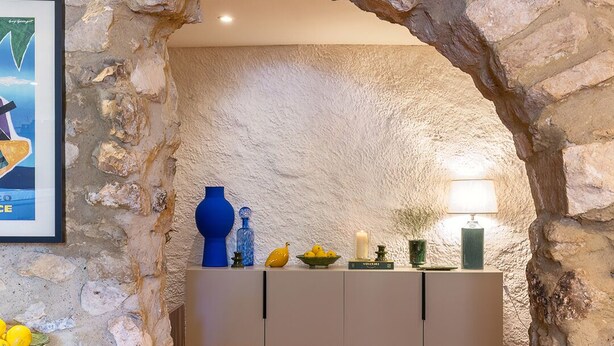 Un arc en pierre mène à une pièce où se trouvent un meuble blanc, un vase bleu, une bougie et une lampe.