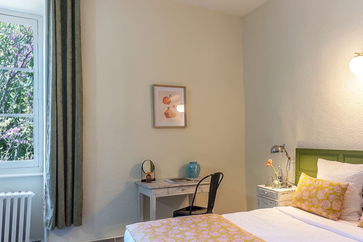 Un dormitorio con cama, escritorio, silla, mesita de noche, lámpara, un cuadro enmarco y una ventana con cortinas.