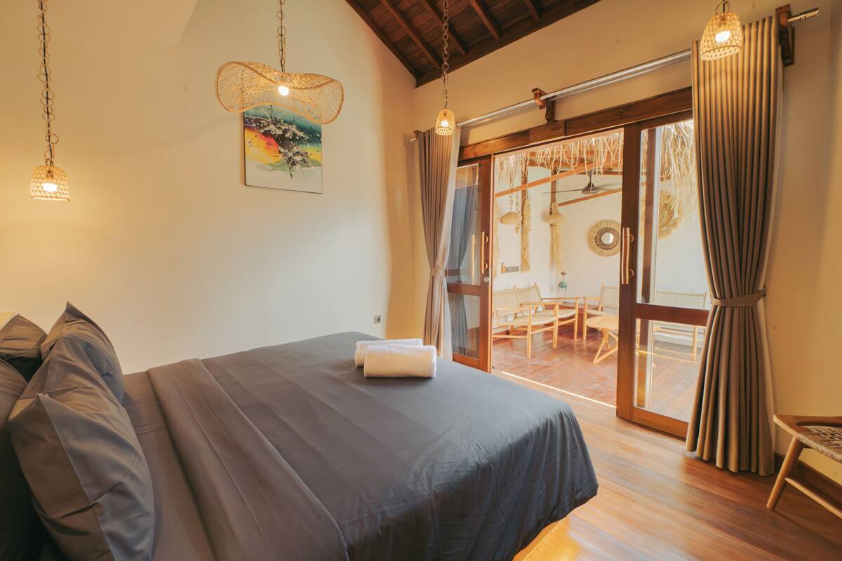 A bedroom with a bed, a hanging light fixture, a painting, and a view of an outdoor area with a table and chairs.