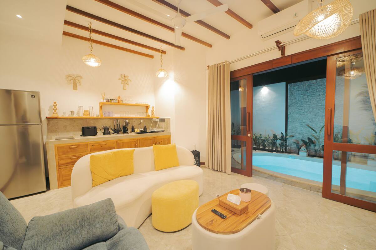 A modern living room with a sofa, ottoman, and coffee table, a kitchen area with a refrigerator, and a large sliding glass door leading to a pool.