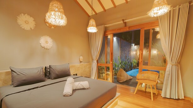 Un dormitorio con cama, almohadas, una toalla enrollada, una silla y vistas a través de un ventanal.