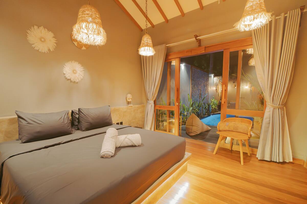 A bedroom with a bed, pillows, a rolled towel, a chair, and a view through a glass door.