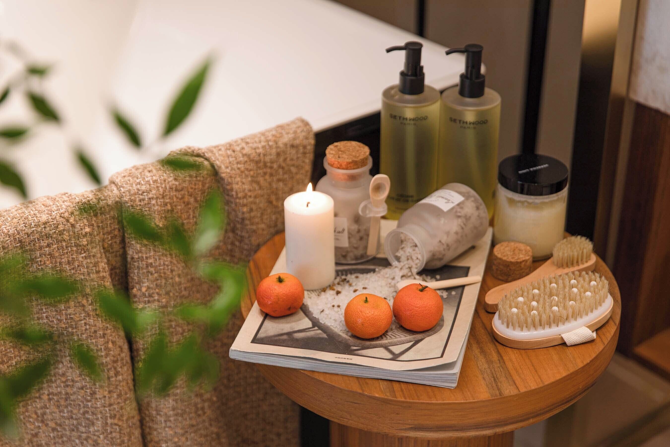 A wooden table with a candle, oranges, a bottle of bath soap, a bottle of lotion, and a hairbrush.