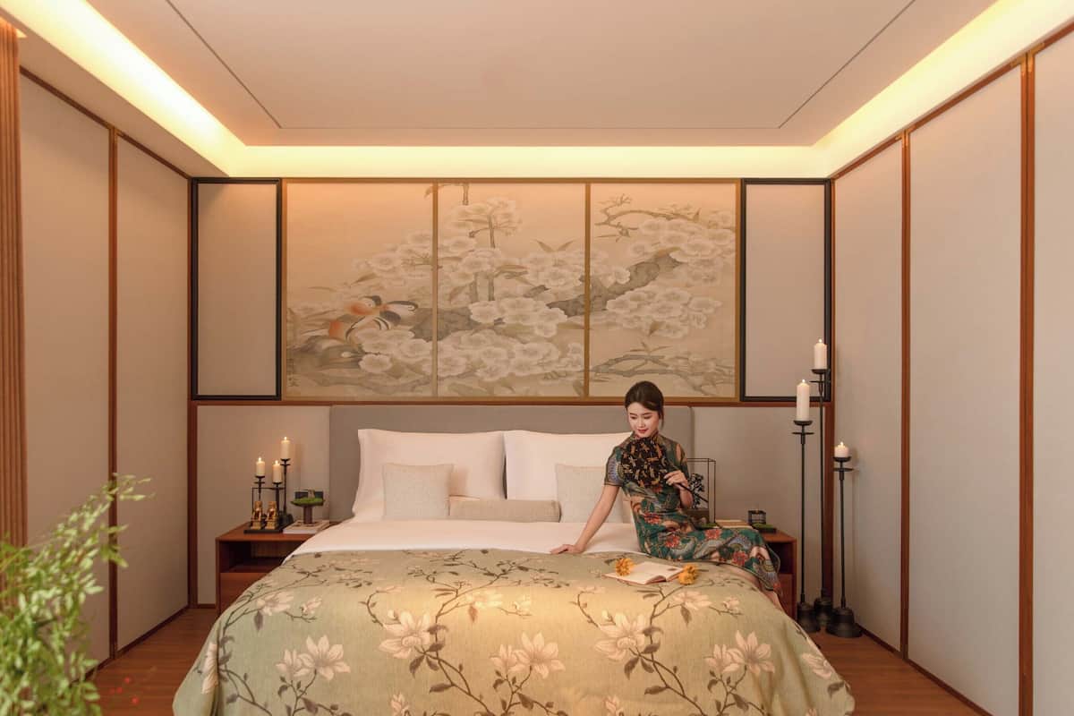 A bedroom with a bed, a nightstand, a lamp, and a wall with a floral painting.
