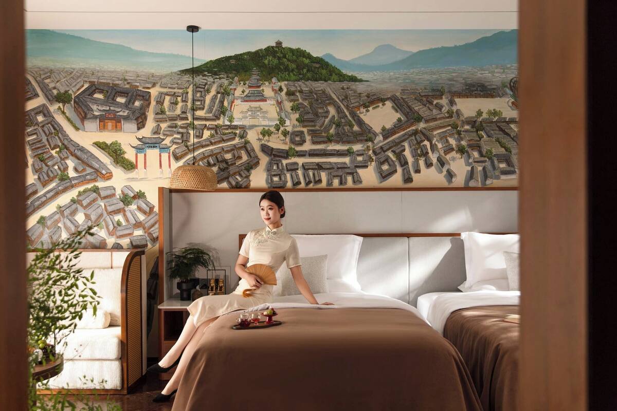 A hotel room with a large mural of a cityscape, a bed with white linens, and a bedside table with a fan and a tray of fruit.