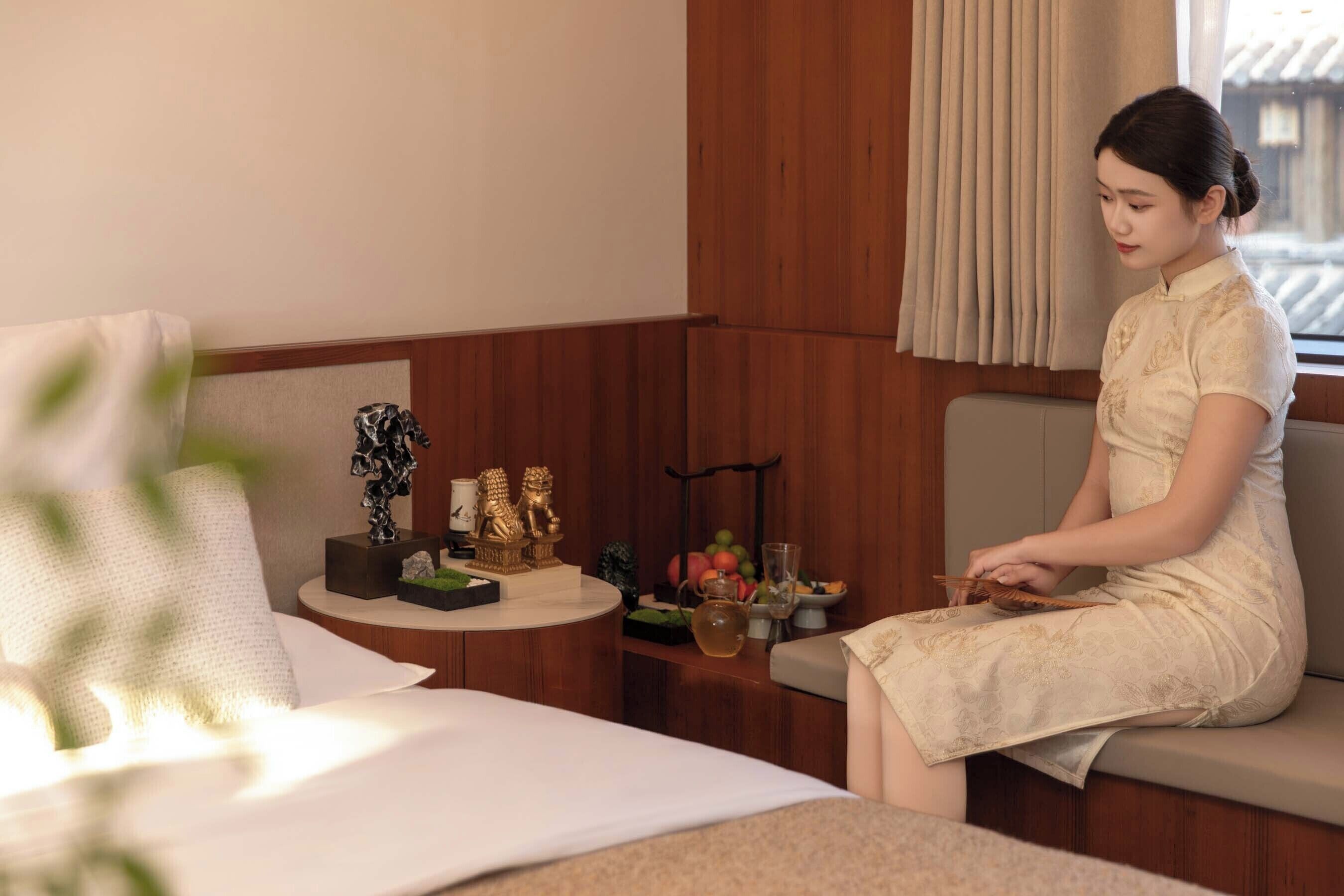 A woman sitting on a sofa in a room with a bed, a small table with decorative items, and a window with curtains.