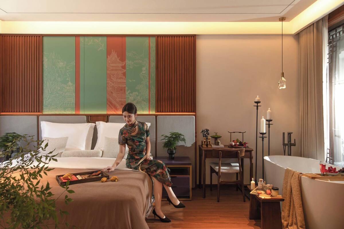 A hotel room with a bed, a desk, a chair, a bedside table, a potted plant, and a large window.
