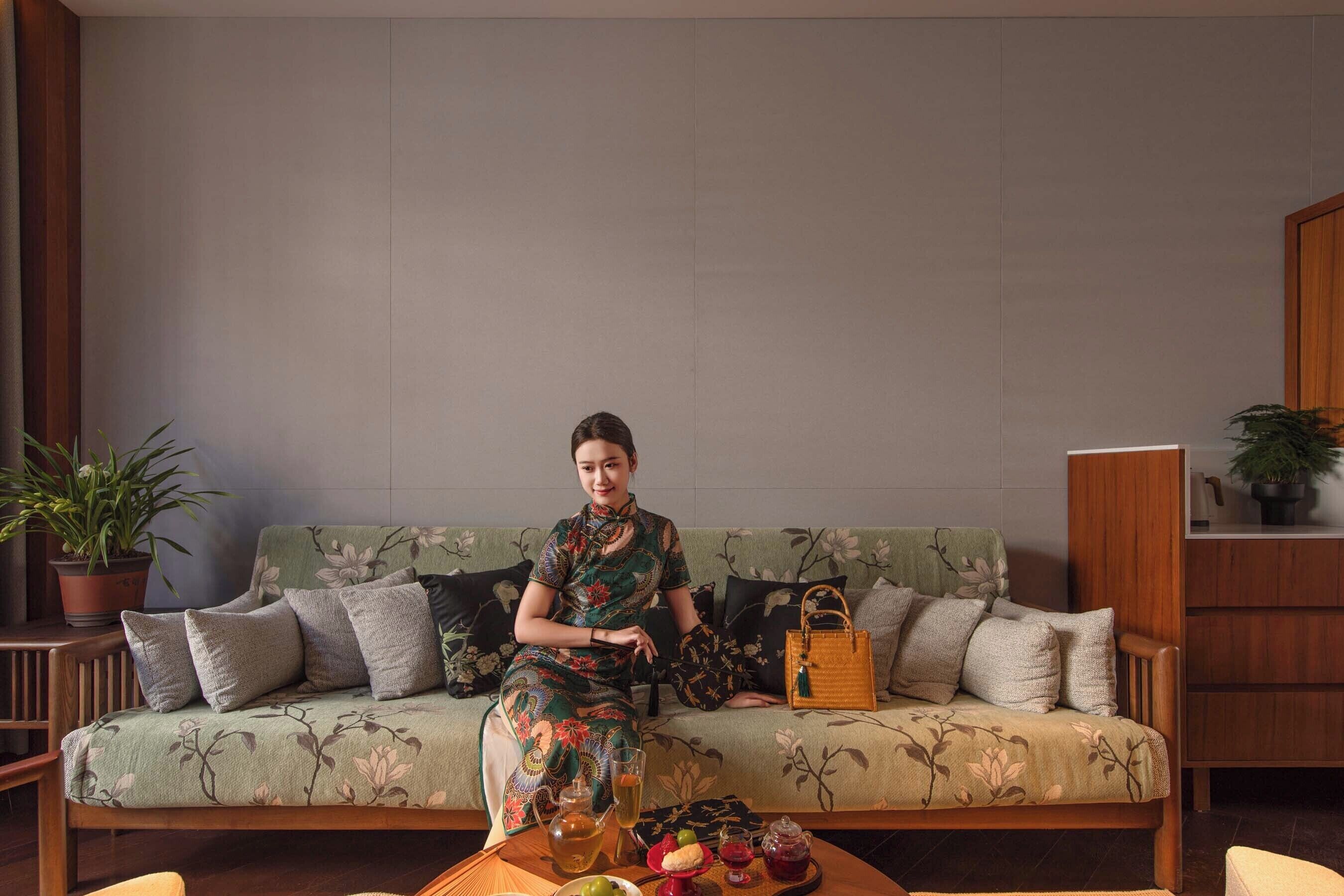 Eine Frau sitzt auf einem Sofa mit floralem Bezug, einem Couchtisch mit Teekanne und Früchten, und einer Topfpflanze links.