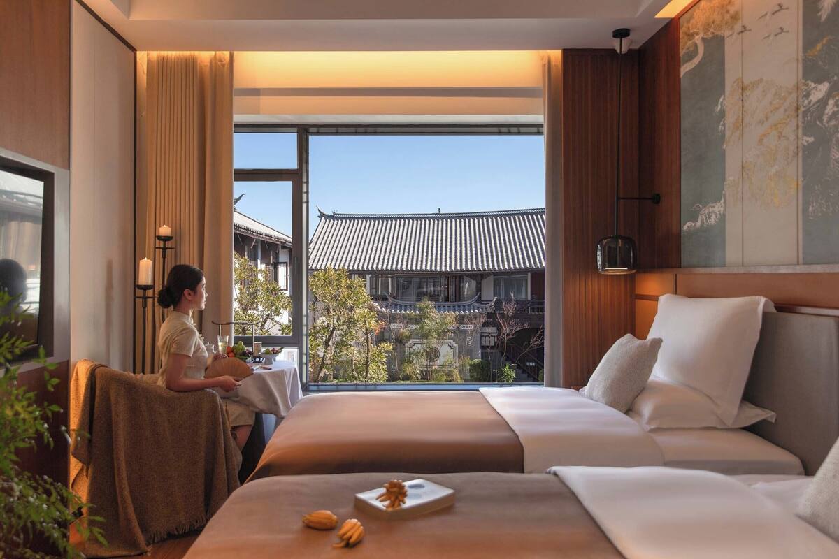 A hotel room with a large bed, a smaller bed, a dining table, and a view of traditional architecture.