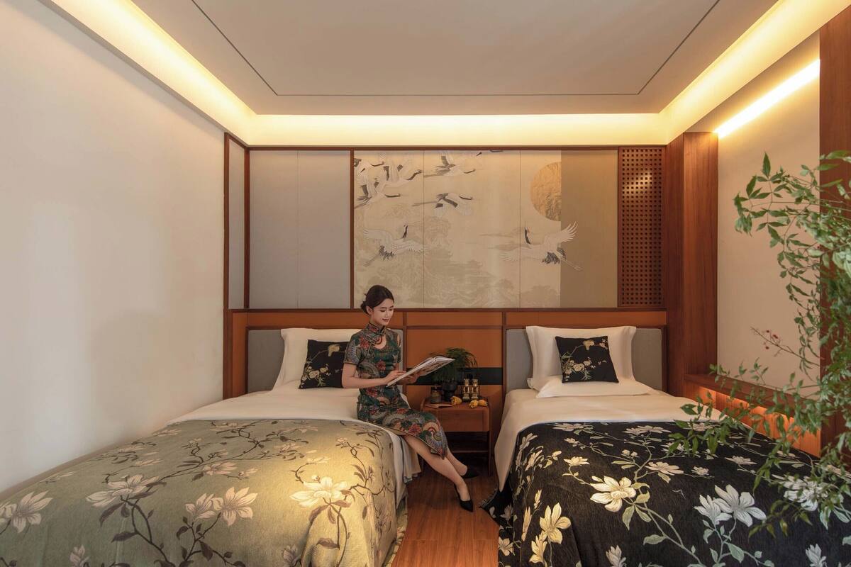 A hotel room with two beds, a wall with a traditional artwork, and a small table with a plant.