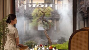 Una persona sentada junto a una ventana con vistas a un edificio tradicional y un bonsái.