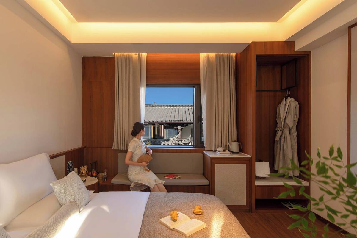 A hotel room with a bed, a seating area, a window with curtains, a robe rack, and a small table with a book and a fan.