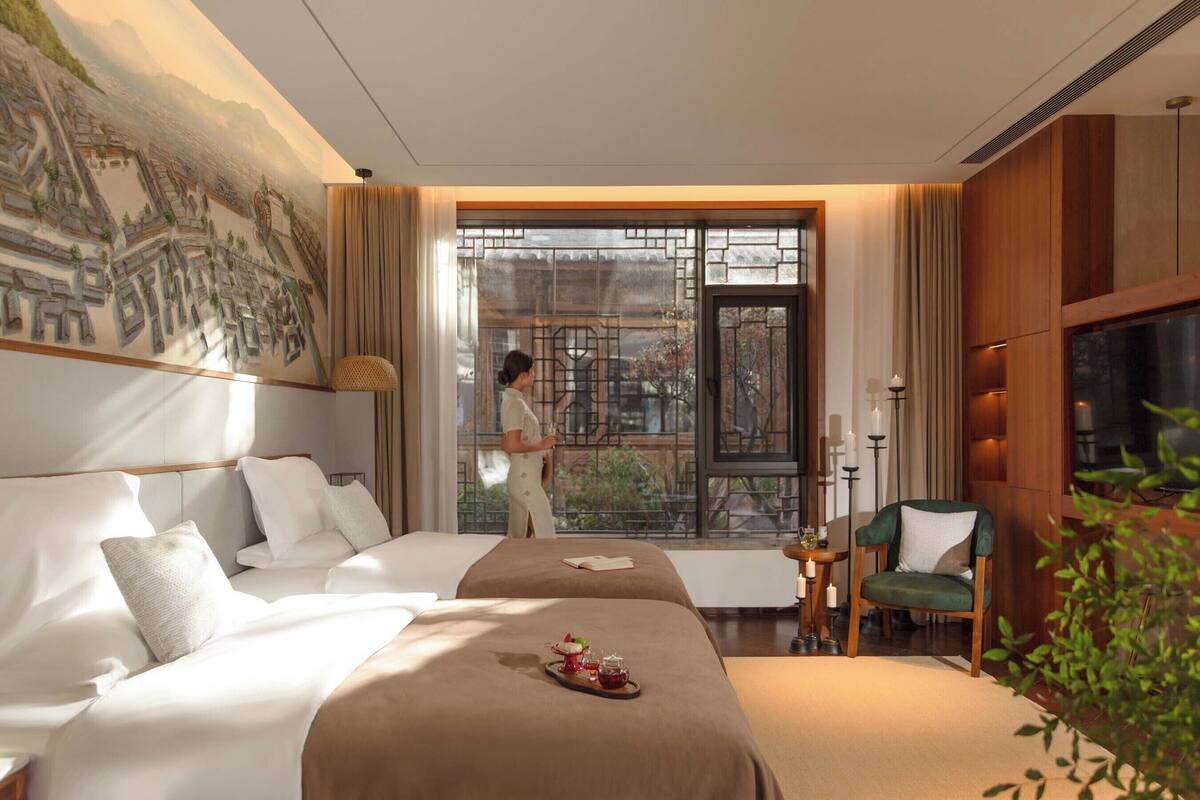 A hotel room with a large bed, a smaller bed, a sitting area with a chair, a table with candles, and a view of a cityscape through a large window.