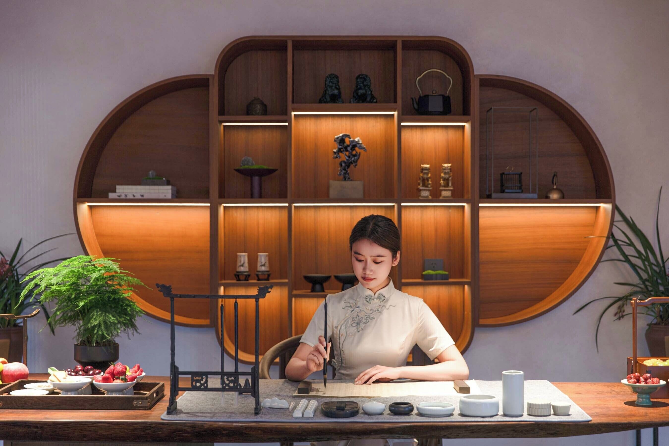Une femme est assise à un bureau en bois et écrit. Une plante et des fruits sont posés à côté d’elle.