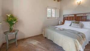Ein Schlafzimmer mit einem Bett, einem Nachttisch mit Blumenvase, einem Fenster und einer Wandleuchte.