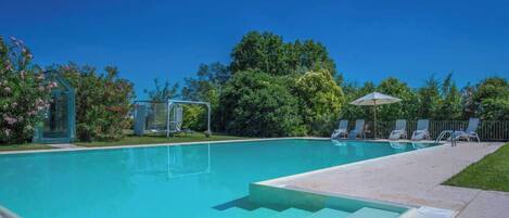 A clear blue swimming pool with steps leading into it, surrounded by lush greenery and a covered seating area with umbrellas.