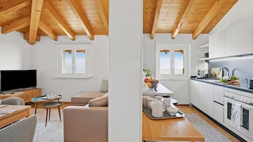 A modern living space with a wooden ceiling, a television, a sofa, a dining area, and a kitchen with white cabinets and appliances.