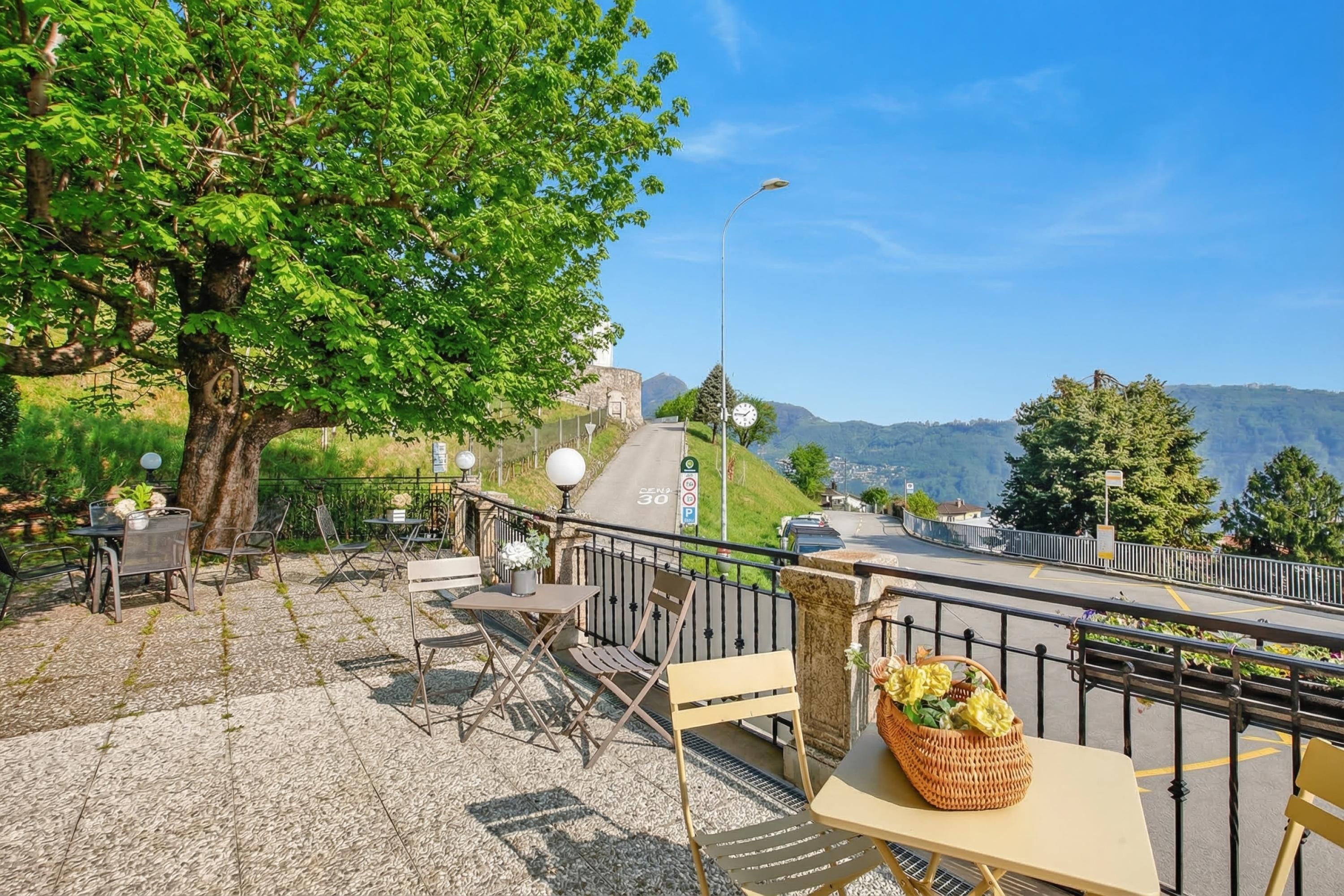 An outdoor seating area with tables and chairs, a tree, and a view of a road and mountains.