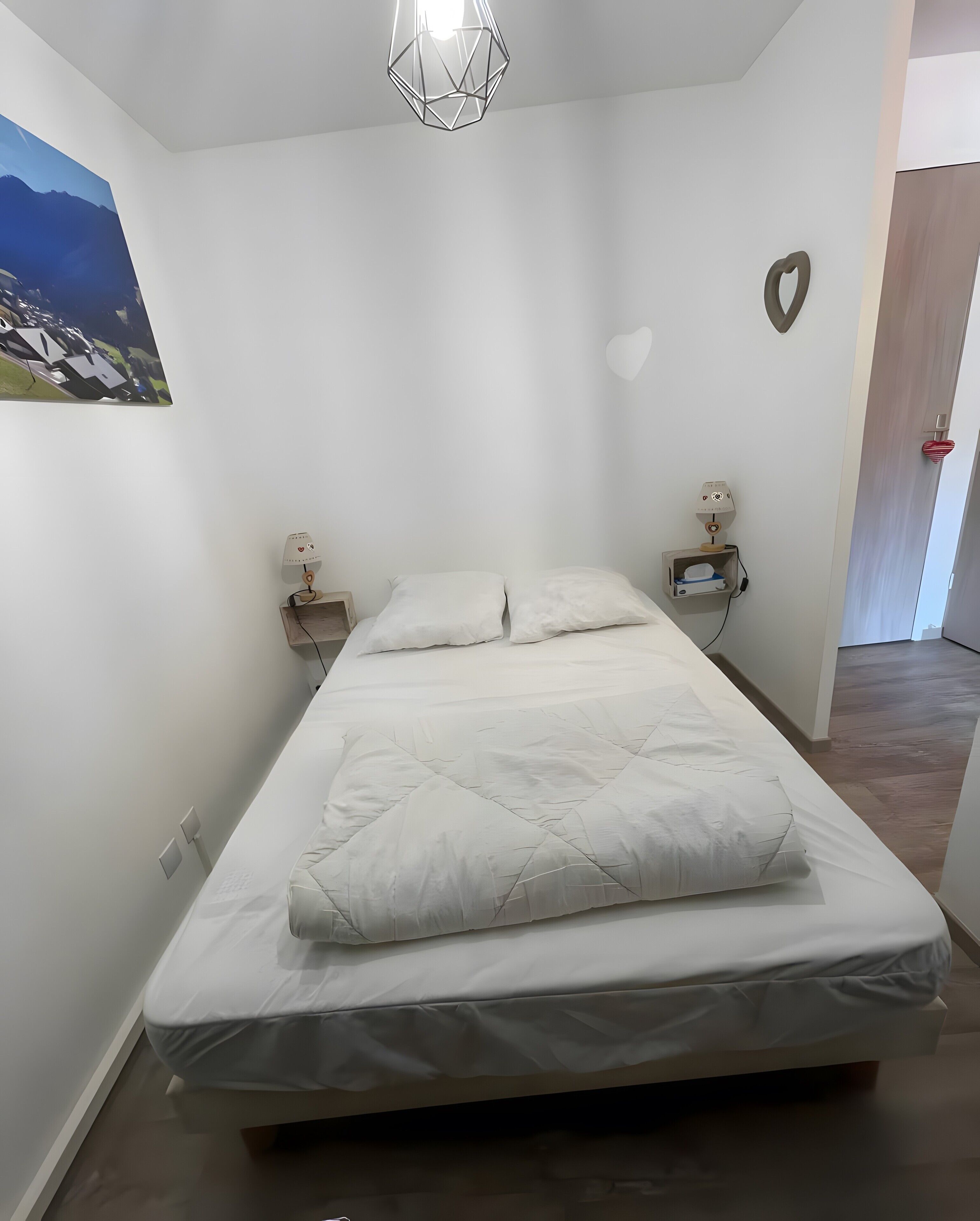 A bedroom with a bed, two bedside tables, a wall-mounted lamp, a heart-shaped decoration, and a painting on the wall.