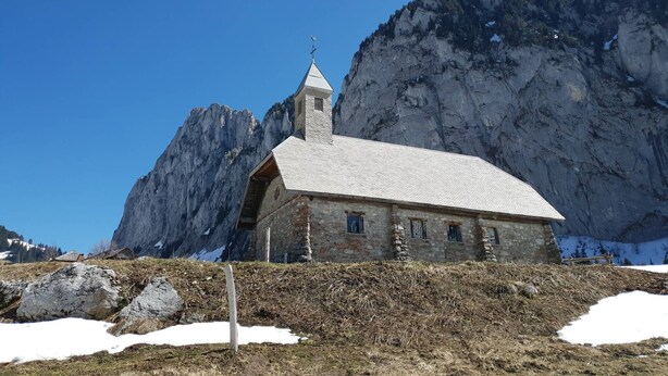 A small stone church with a bell tower, set against a mountainous backdrop.