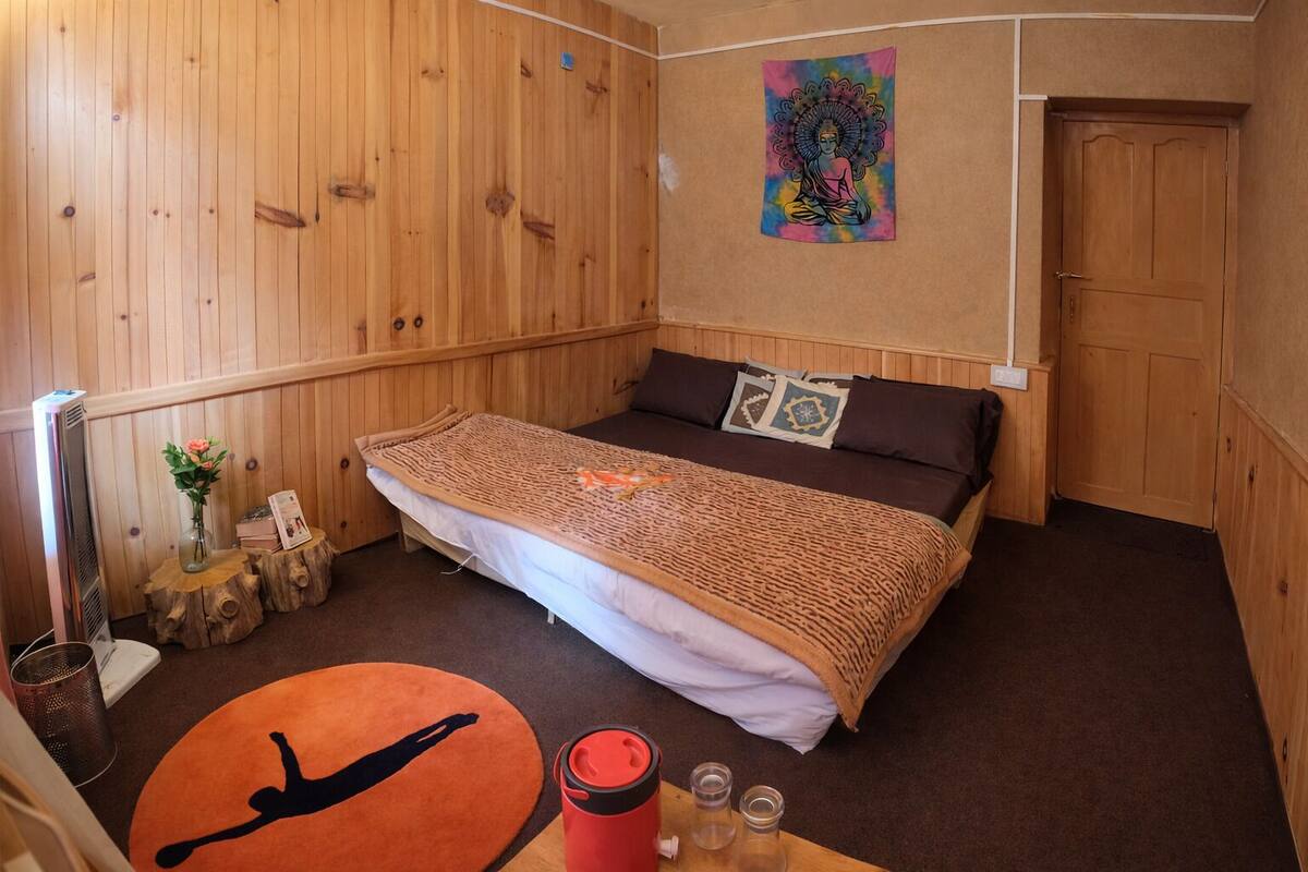 A wooden-paneled room with a bed, a wall tapestry, and a small table with a red water bottle and glasses.
