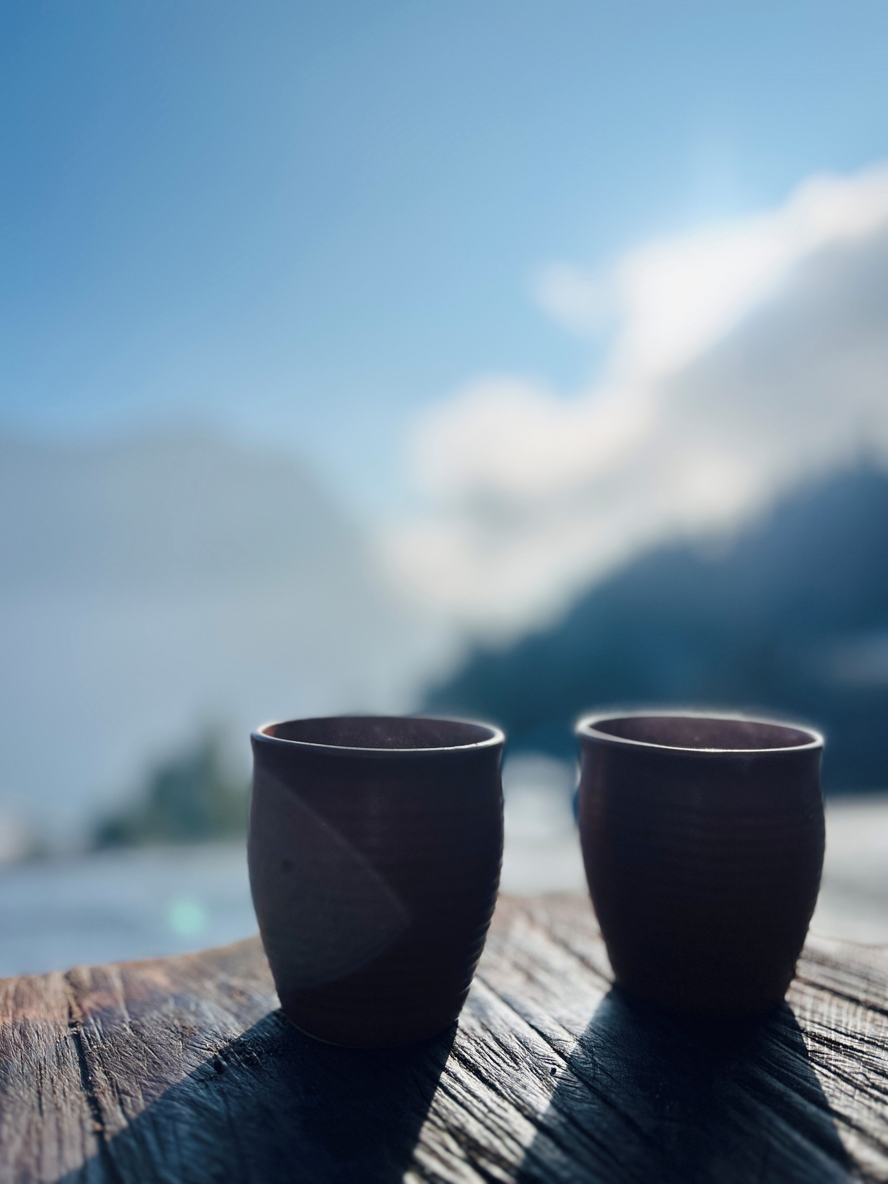 Dos tazas de cerámica sobre una superficie de madera, con un fondo desenfocado de montañas y cielo.