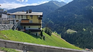 A multi-story building with a staircase leading to the entrance, surrounded by lush greenery and mountains in the background. - Cozy Apartment With a Beautiful View (Kappl)