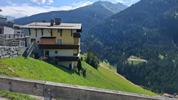 Un edificio de varios pisos con una escalera que conduce a la entrada, rodeado de abundante vegetación y montañas al fondo.