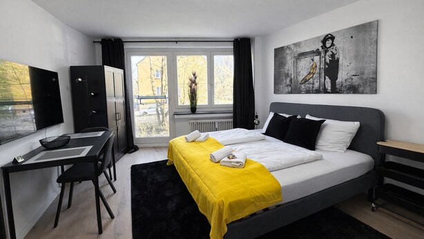 Moderni hotellihuone, jossa on sänky, työpöytä, tuoli, patteri ja ikkuna, josta on näkymä ulos.