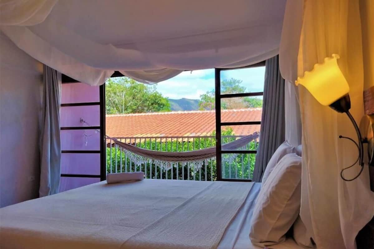 Quarto com cama, rede e vista para um prédio com telhado vermelho através de grandes janelas.