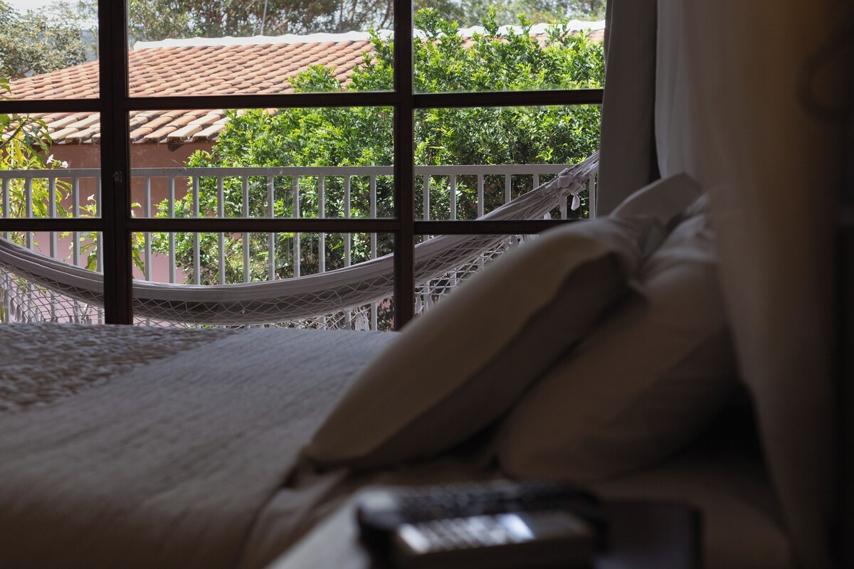 A bedroom with a bed, a remote control, and a view of a balcony with a railing and greenery.