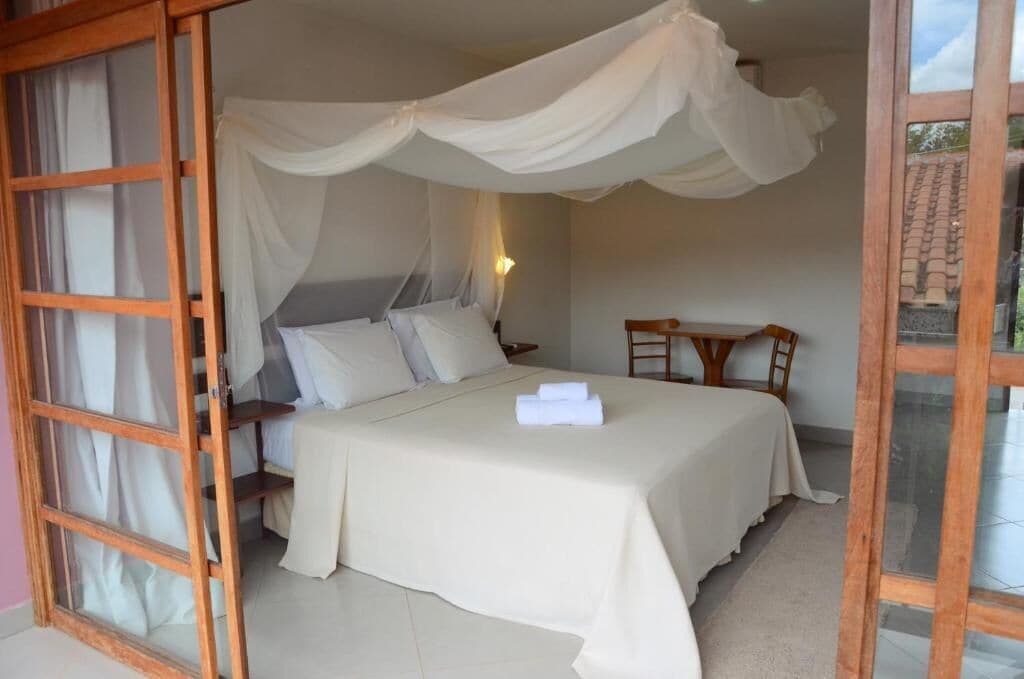 A bedroom with a canopy bed, a wooden chair, a small table, and a window with curtains.