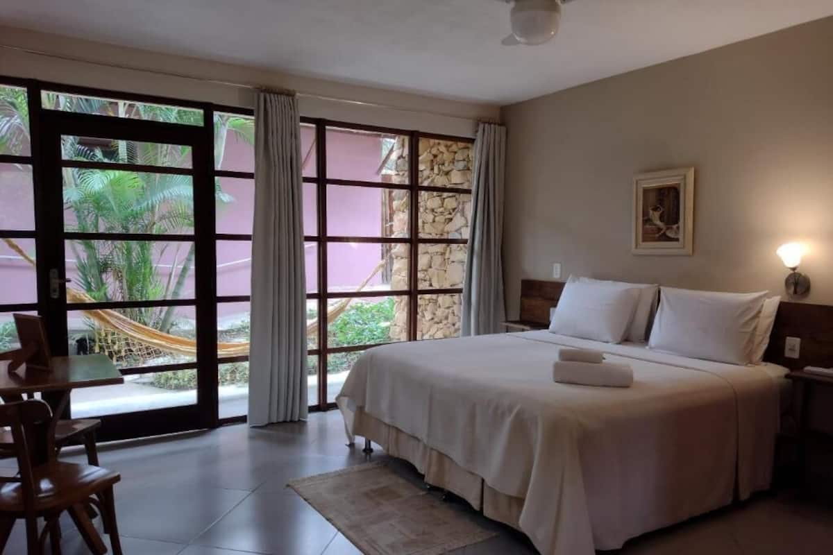 Quarto com cama, escrivaninha, cadeira, janela com cortinas e vista para o verde lá fora.