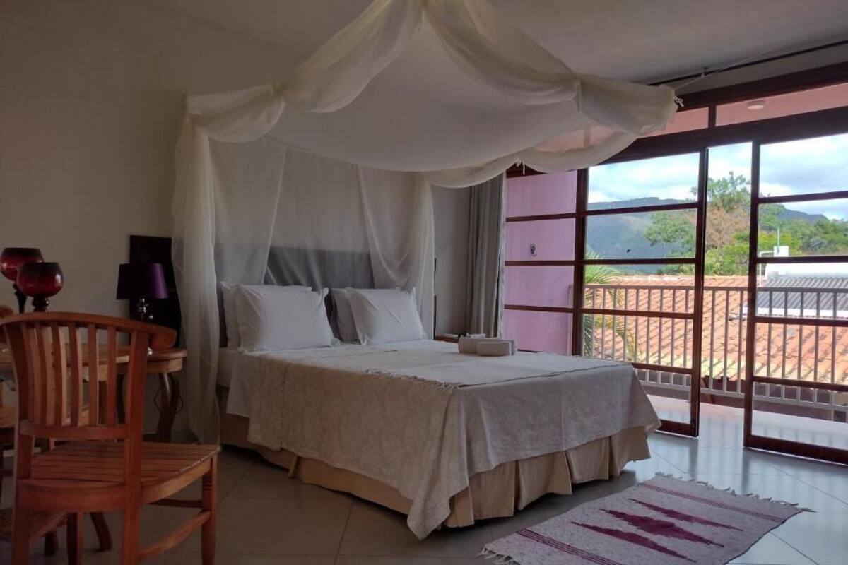 Quarto com cama, cadeira, mesa de cabeceira, luminária e vista para o exterior por grandes janelas.