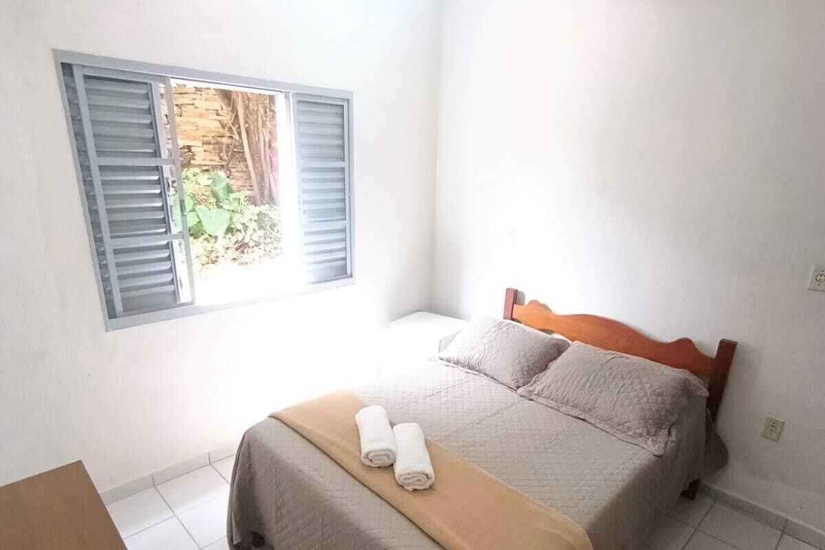 Un dormitorio con cama, ventana con contraventanas, una mesita y una alfombra.