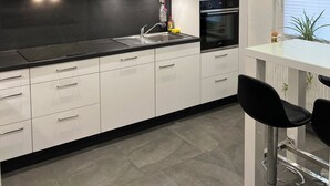 A modern kitchen with white cabinets, a black countertop, a stainless steel range hood, a built-in oven, and a bar area with two black bar stools. - Andy's apartment (Malsch)