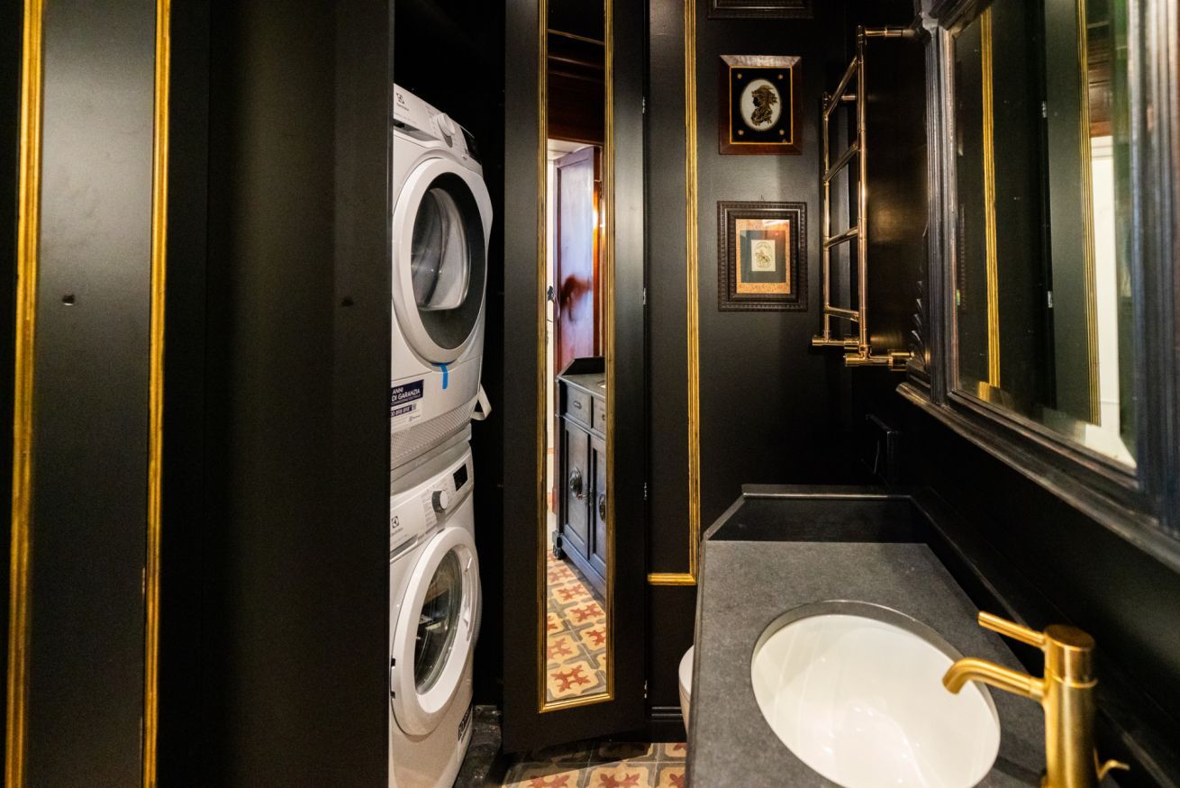 Een kleine hotelbadkamer met een wasmachine, een wastafel met gouden kraan en kunstwerken in lijsten aan de muur.