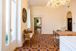 Ein Zimmer mit einem gemusterten Boden, einem Esstisch, Stühlen, einer Kronleuchter und Blick durch eine offene Tür.